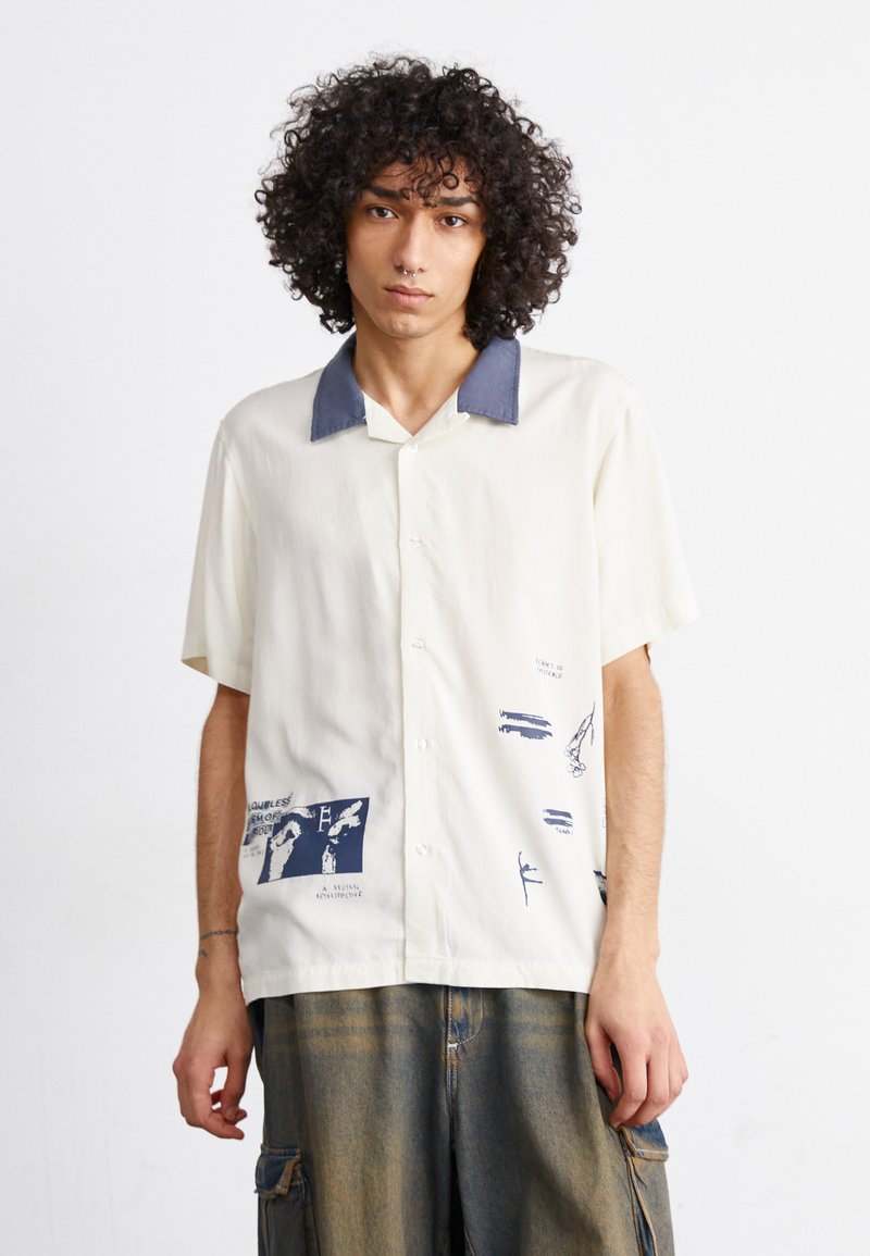 Camicia a maniche corte color crema con colletto blu navy. Presenta grafiche stampate in blu, tra cui disegni astratti e testo, realizzata in tessuto leggero.