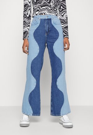 High-waisted flared jeans with light and dark blue wavy denim panels paired with white platform sneakers and a black-and-white patterned top.