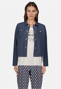 Femme portant une veste boutonnée bleu marine sur un T-shirt imprimé blanc et un pantalon à motifs bleu marine, se tenant devant un fond uni.