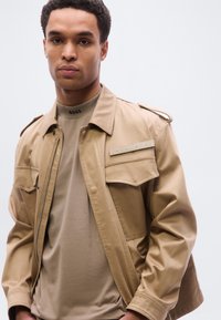 Μπεζ βαμβακερό μπουφάν με φερμουάρ και επωμίδες στους ώμους, συνδυασμένο με μπεζ εφαρμοστό T-shirt με λογότυπο στο γιακά.