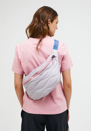 Frau trägt ein hellrosa T-Shirt und eine gesteppte graue Umhängetasche mit rosa Reißverschluss und blauem Trageriemen über dem Rücken.