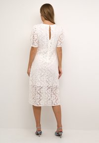 White lace midi dress with short sleeves, floral pattern, keyhole back detail, and fitted waist. Worn with light blue high-heeled shoes.