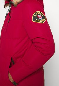 Czerwona pikowana parka z kapturem wykończonym futrem, z czarno-żółtą naszywką z logo na rękawie i bocznymi kieszeniami na zamek. Gładka faktura.