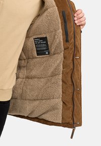 Bruine corduroy jas met een crème kleurige fleece voering, voorzien van een zijrits, gewatteerde textuur en een verzorgingslabel zichtbaar aan de binnenkant.