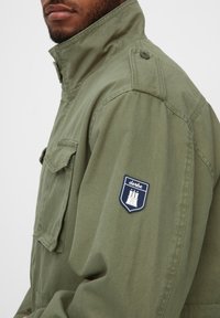 Olivengrøn jakke med høj krave, knapdetaljer på skulderne og en lomme på venstre bryst. Har et blåt skjoldpatch med et slotdesign.