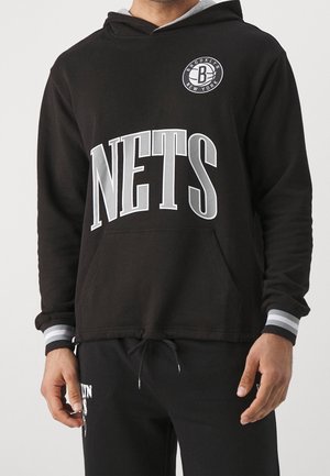 Muž, který má na sobě černou mikinu s bílým nápisem "NETS" a logem Brooklyn New York na prsou, v kombinaci s černými kalhotami.