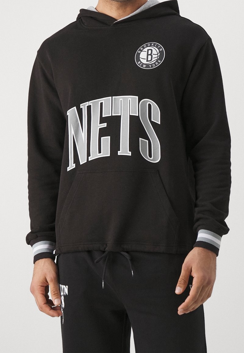 Homme portant un sweat à capuche noir avec le texte blanc "NETS" et un logo Brooklyn New York sur la poitrine, assorti à un pantalon noir.