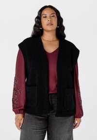 Zwarte, gemarmerde mouwloze vest met twee voorkruikrings, gedragen over een bordeaux langemouwen top met bloemenborduursel, gecombineerd met donkergrijze spijkerbroek.