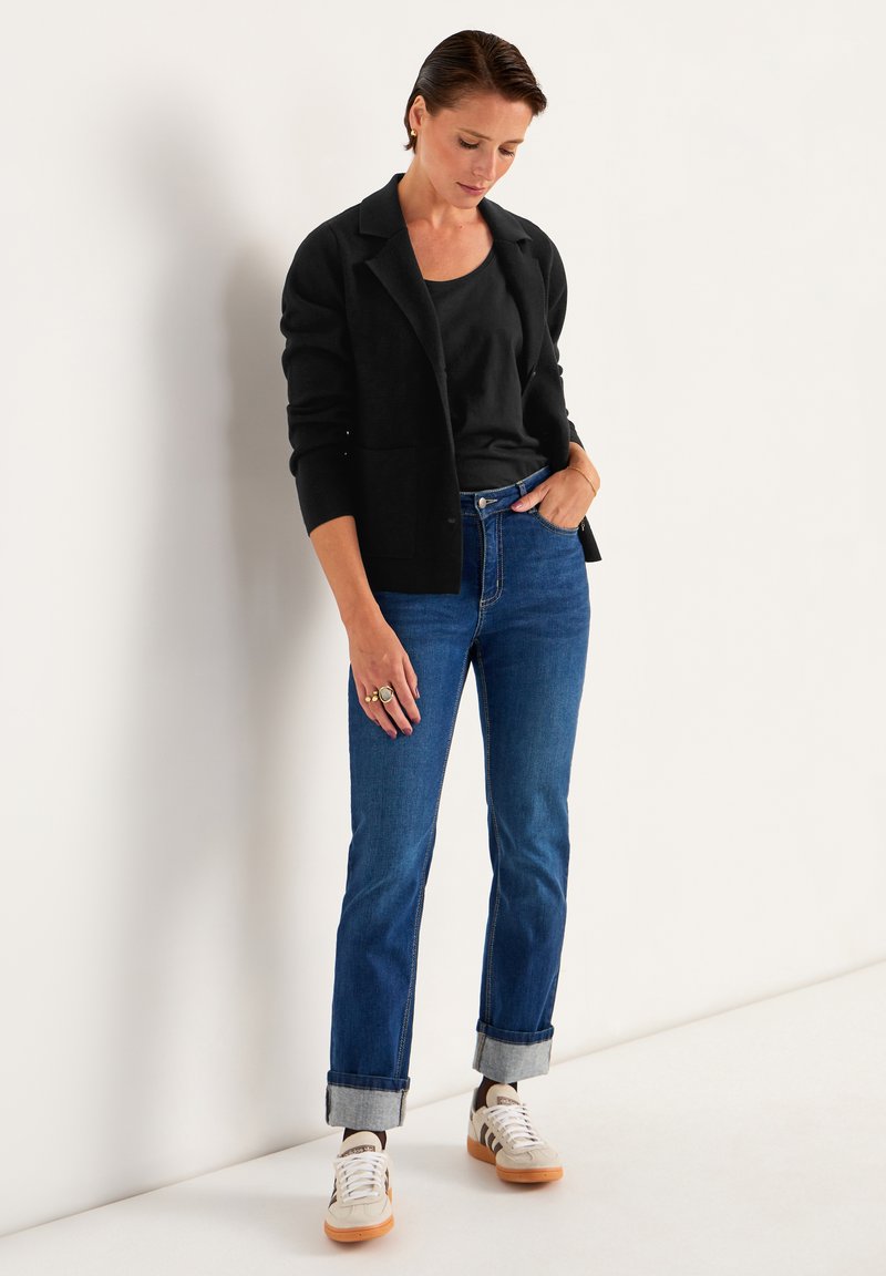 Sort cardigan, sort tanktop, blå lige ben jeans med opsmøg, og hvide sneakers med brune detaljer. Enkle smykker, afslappet positur.