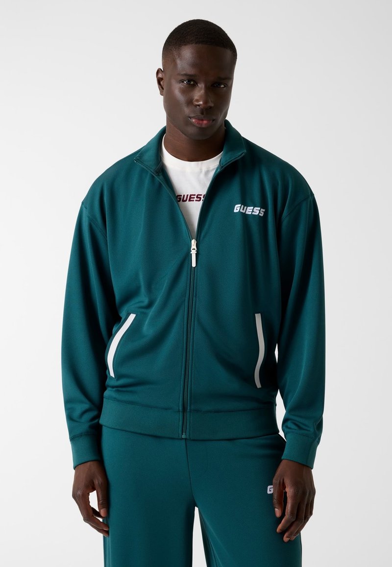 Turkooise trainingsjack met ritssluiting, witte accenten en zijzakken; uitgevoerd in een gestructureerde stof en met het merklogo op de borst.