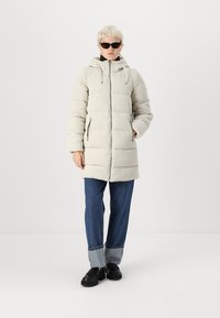 Doudoune beige clair avec un design matelassé, capuche, fermeture éclair à l'avant, et deux poches zippées sur les côtés ; associée à un jean bleu ample et des chaussures noires.