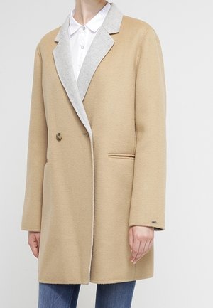 Woman wearing a beige wool coat with gray lapels over a white buttoned shirt and blue jeans, standing against a plain background.