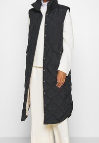 Schwarze gesteppte lange Weste mit hohem Kragen, Druckknöpfen und seitlichen Taschen, aus leichtem Stoff gefertigt, über einem cremfarbenen Pullover und Hose getragen.