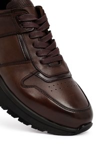 Bruine leren sneaker met een gestructureerde bovenkant, zes vetersluitingen, perforaties en een zwarte rubberen zool. Glad oppervlak met contrasterende stiksels.