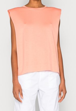 Mouwloos perzikkleurig topje met gevoerde schouders, gedragen met witte broek, getoond op een persoon tegen een effen lichte achtergrond.