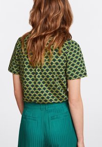 Blouse verte avec un motif floral en marine et jaune. Manches courtes et coupe décontractée. Portée avec un pantalon plissé teal taille haute.