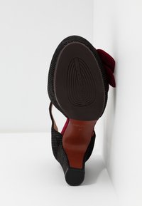 Chaussure à talon haut noire avec petits points blancs, détail nœud rouge et semelle marron lisse, inclinée contre un fond blanc en angle.