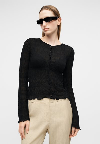 Γυναίκα με μαύρο υφασμάτινο τοπ με κουμπιά και μακριά μανίκια, μπεζ παντελόνι ψηλόμεσο και στενά μαύρα γυαλιά ηλίου, μπροστά σε απλό φόντο.