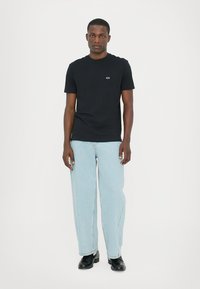 Svart t-shirt med kort ärm och en liten logotyp vid bröstet, tillsammans med ljusblå, löst sittande jeans och svarta skor. Enkel, avslappnad design.