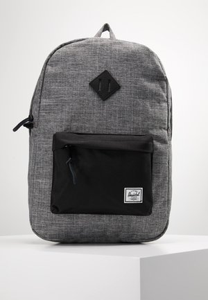 Zaino in tessuto grigio con tasca frontale nera e toppa a forma di diamante su una superficie bianca.