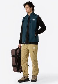 Veste sans manches bleue sur chemise noire à manches longues, pantalon kaki, sac polochon marron et bottes foncées ; présente des accents de logo et des fermetures éclair.