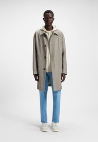 Cappotto grigio a quadri con una vestibilità comoda, grandi bottoni e spacchi laterali, indossato sopra un leggero felpa con cappuccio e abbinato a jeans azzurri chiari e sneakers.