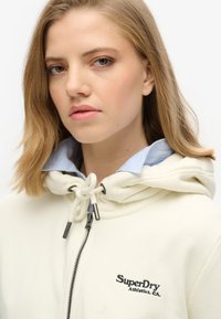 Sudadera crema con capucha y cuello a rayas azules. Cuenta con un cierre frontal y el logo bordado "SuperDry Athletics, CA" en el pecho.