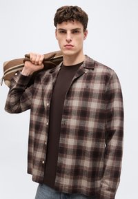 Νεαρός άνδρας που φορά καφέ πουκάμισο με καρό σχέδιο και σκουρόχρωμο μπλουζάκι, κρατώντας μια καφέ τσάντα στον ώμο του με φόντο μια απλή επιφάνεια.