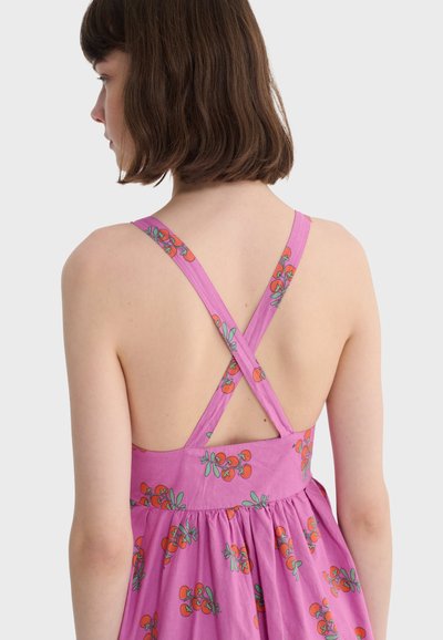 Jeune femme aux cheveux châtain coupés au carré, portant une robe rose sans manches avec un imprimé cerises rouges et des bretelles croisées dans le dos, vue de dos.