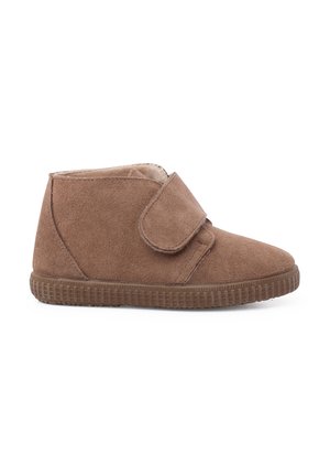 Zapato de ante marrón estilo pantufla hasta el tobillo con correa de velcro y suela de goma texturizada, mostrado desde el lado exterior sobre un fondo blanco.