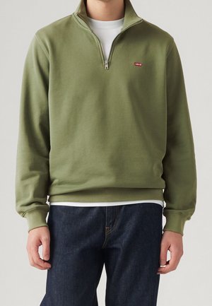Personne portant un sweat-shirt vert olive à fermeture éclair quart, par-dessus une chemise blanche et un jean bleu foncé, debout devant un fond uni.