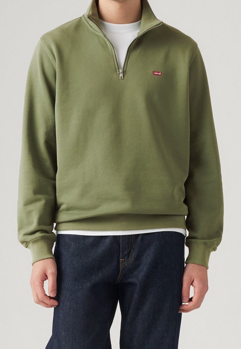 Personne portant un sweat-shirt vert olive à fermeture éclair quart, par-dessus une chemise blanche et un jean bleu foncé, debout devant un fond uni.