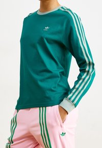 Teal långärmad skjorta med grå krage och tre ljusgrå ränder på ärmarna. Vit logotyp på bröstet. Bomullstyg, figurnära design.