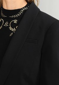 Černý blazer se strukturovanými klopami, se malou náprsní kapsou. Pod ním je černý top s zlatým květinovým vyšíváním a vysokým výstřihem.