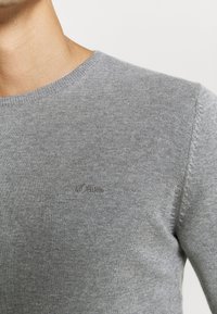 Grauer Strickpullover mit Rundhalsausschnitt, der dezente Rippstrickdetails und ein kleines gesticktes Logo auf der Brust aufweist. Angenehme, glatte Textur.