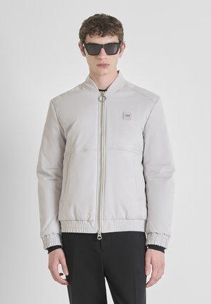 Jeune homme portant une veste bomber grise clair avec une fermeture éclair en anneau et des lunettes de soleil noires, se tenant devant un fond blanc uni.