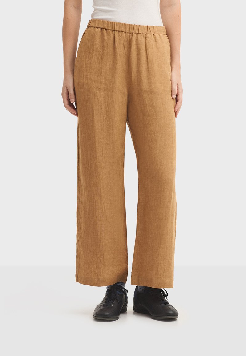 Pantalon en lin marron à jambes larges avec une ceinture élastique, présentant un tissu texturé et une coupe ample, associé à des chaussures noires.
