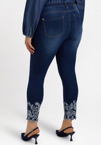 Navy siniset farkut, joissa on sovitettu malli, ja joissa on valkoista paisley-kirjontaa lahkeiden päällä sekä kaksi takataskua. Teksturoitu farkkukangas.