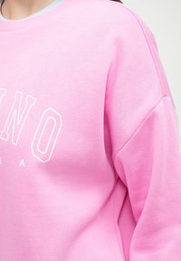 Sweat-shirt rose avec texte blanc. Présente un col côtelé et une coupe loose, fabriqué en tissu doux avec une texture lisse.