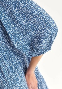 Blaues Kleid mit weißem Blumenmuster, das über puffige Ärmel verfügt. Der Stoff ist leicht und strukturiert, mit gerafften Details an den Ärmeln.