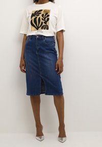 Camiseta blanca con estampado botánico en negro y dorado, combinada con una falda lápiz de mezclilla oscura con una abertura frontal y ligero deshilachado en el dobladillo.