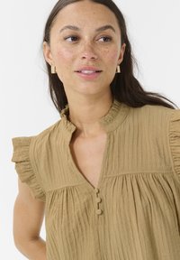 Camisa caqui con mangas acampanadas, material texturizado y tapeta de botones. Presenta un cuello mao y tejido recogido debajo del busto.
