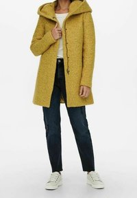 Gelber Wollmischmantel mit Kapuze, Knopfverschluss und strukturierter Oberfläche, kombiniert mit dunklen Denim-Jeans und weißen Sneakern.