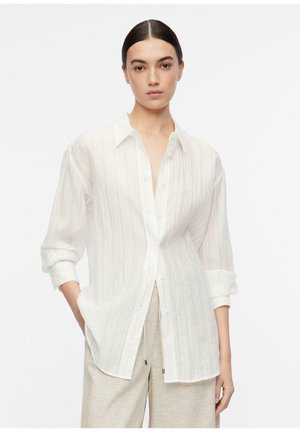 Woman with dark hair in a white textured button-up shirt with rolled sleeves and beige wide-leg pants, standing against a white background.