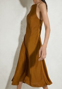 Kobieta w bezrękawnikowej sukience midi w kolorze rdzy, stojąca przy fakturowanej, off-white ścianie, ukazująca widok z boku i częściowo z przodu.