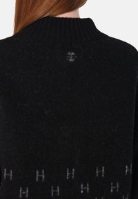 Svart stickad tröja med ribbad krage, med en slät textur och ett ljusgrått logomönster i nederkant. Liten dragning synlig på baksidan.