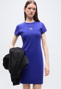 Γυναίκα με μπλε φόρεμα με κοντά μανίκια και μήκος μέχρι το γόνατο, με το λογότυπο "CK Calvin Klein", κρατώντας ένα μαύρο σακάκι στο αριστερό της χέρι.
