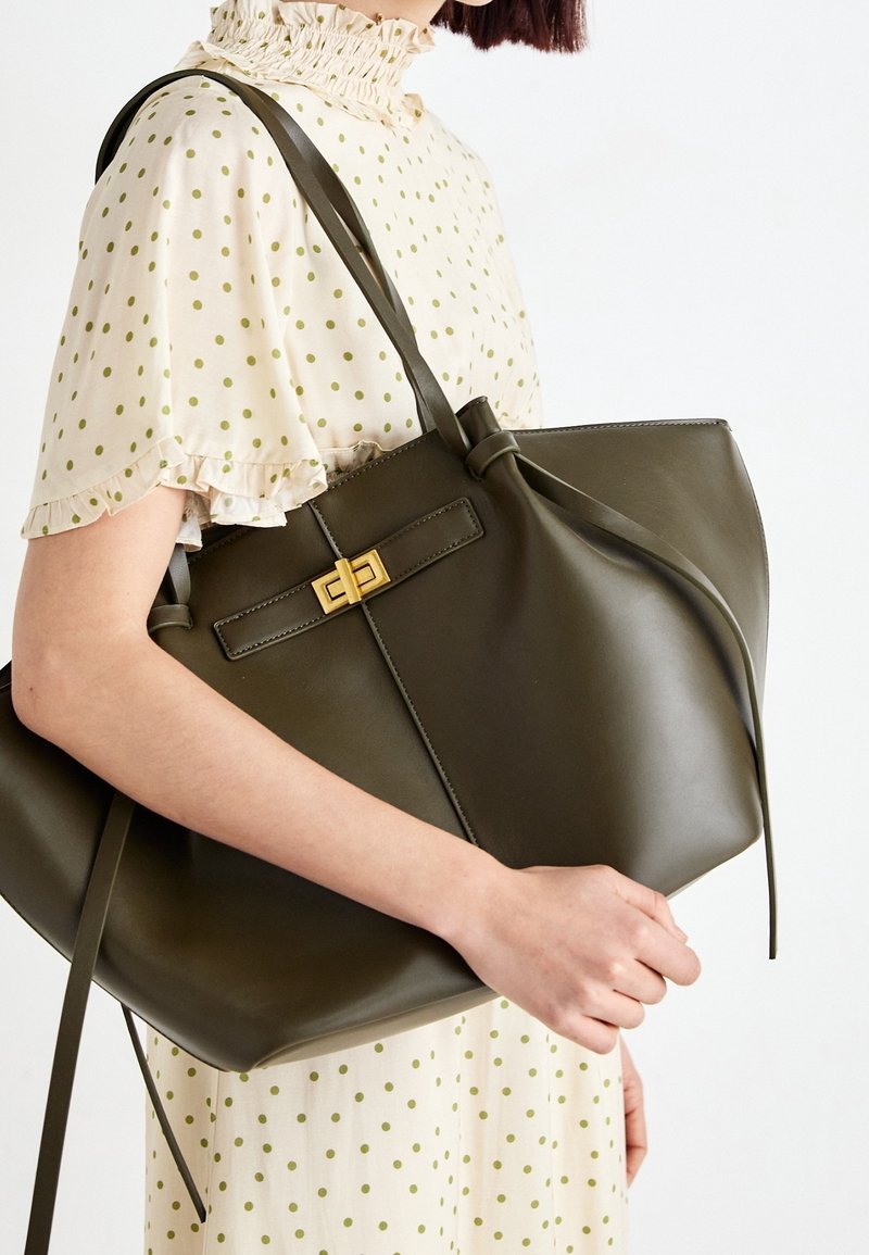 Oliivinvihreä nahkainen olkalaukku, jossa on litteä pohja ja pehmeä, jäsennelty muoto. Varustettu keskellä olevalla rusettiyksityiskohdalla ja kullanvärisellä lukolla.
