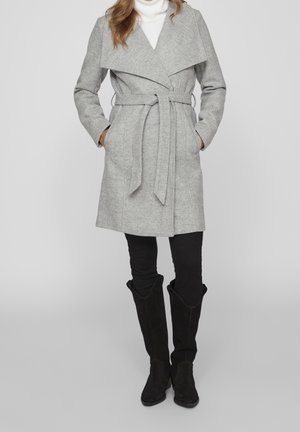 Vrouw draagt een grijze wollen jas met riem over een witte coltrui, zwarte broek en zwarte kniehoge laarzen, staand met handen in de zakken.