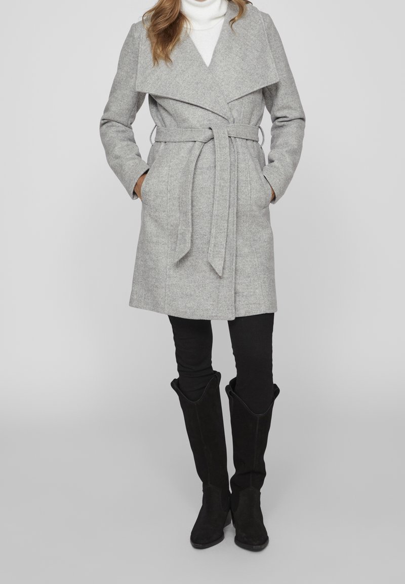 Femme portant un manteau en laine gris ceinturé par-dessus un col roulé blanc, un pantalon noir et des bottes noires montantes jusqu'aux genoux, debout les mains dans les poches.